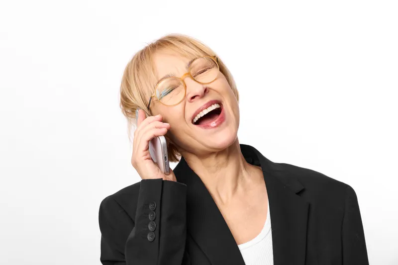 Laughing woman with glasses speaking on a mobile phone, wearing a black blazer. Studio portrait on a clean white background, business communication and phone call in office style.