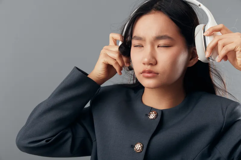 Young woman wearing headphones with closed eyes, enjoying music or sound in quiet environment. Portrait of female with long black hair and dark clothing on plain background.