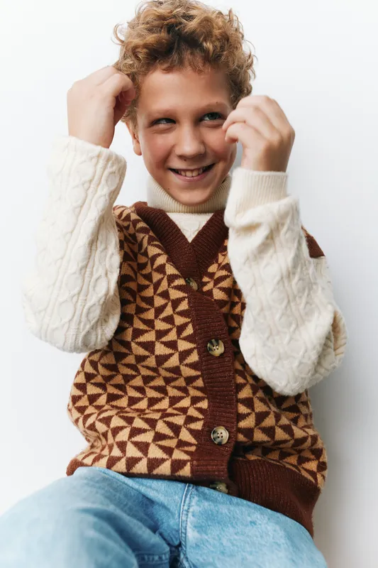 A cheerful young boy with curly hair wears a patterned knit vest over a cream sweater, smiling while adjusting his outfit in a bright studio setting.