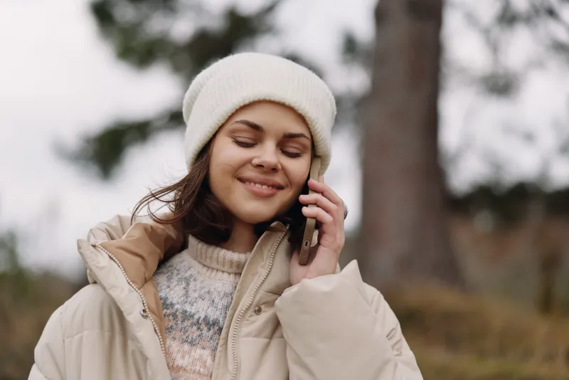 Woman on a chilly outdoor day speaks on a phone, wearing a warm coat and beanie, smiling softly as trees blur in the background with autumnal air.