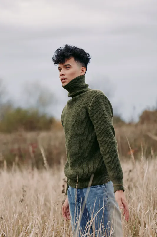 A genuine outdoor portrait of a young man wearing a green turtleneck sweater standing in tall grass, captured with realistic lighting and natural expression conveying authenticity.