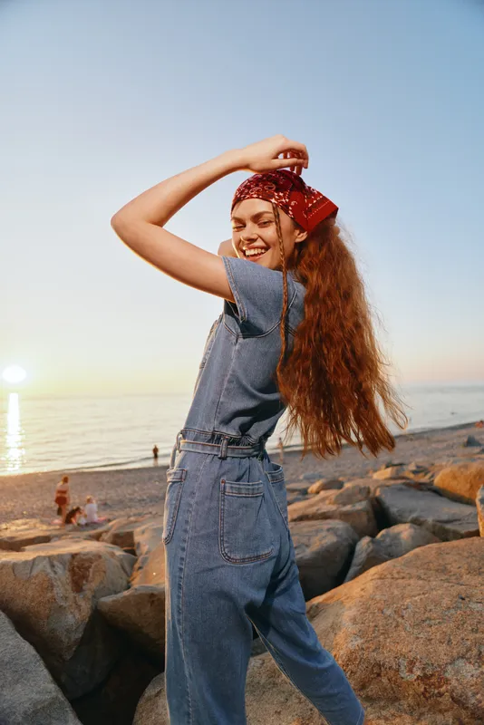 smiling woman in denim jumpsuit with red bandana enjoying sunset by the rocky beach with calm sea and clear sky background