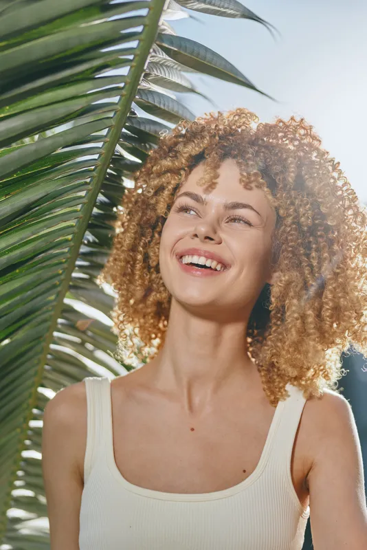 smiling woman with curly hair enjoys sunny day near tropical palm leaves, wearing sleeveless top, natural light highlighting joyful expression and summer mood.