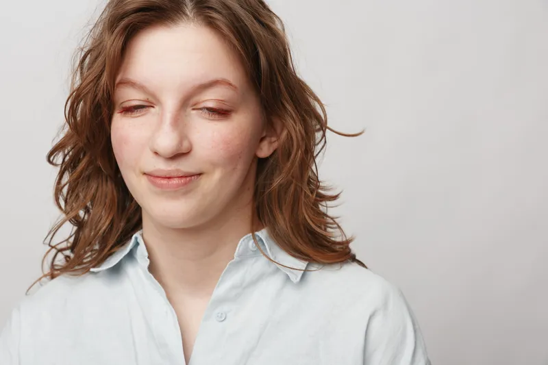 Young woman with closed eyes and slight smile wears light blue shirt against plain background. Concept of calm, relaxation, peaceful moment, and natural beauty in casual style.