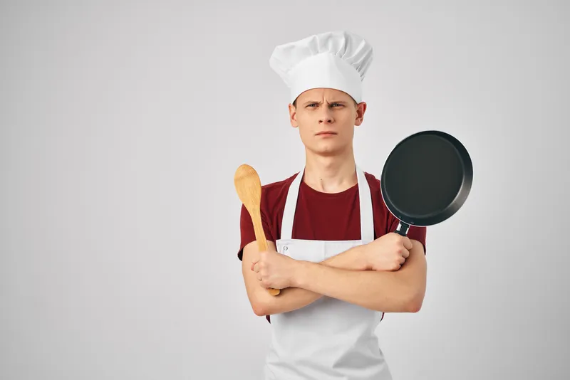 Мужчина в белом фартуке держит в руках сковороду, готовя еду для профессионалов на светлом фоне