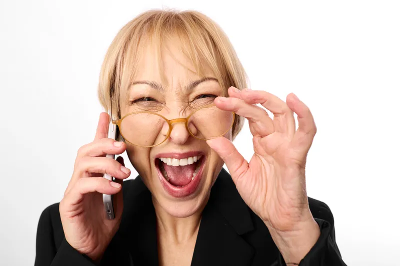 Happy senior woman, smiling widely while holding smartphone to ear, adjusting eyeglasses with one hand, laughing expression, studio portrait, bright white background, close up