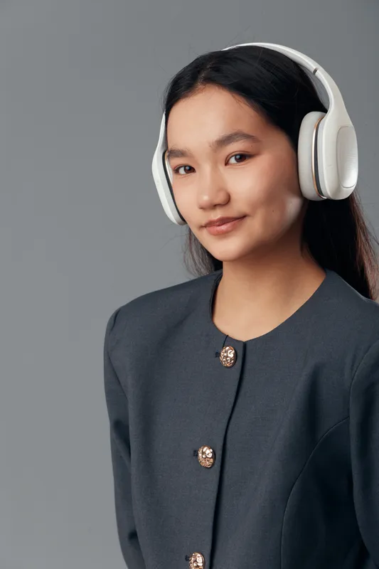 Young woman with long black hair wearing white headphones and a dark jacket with buttons posing on gray background with slight smile and confident expression indoors.
