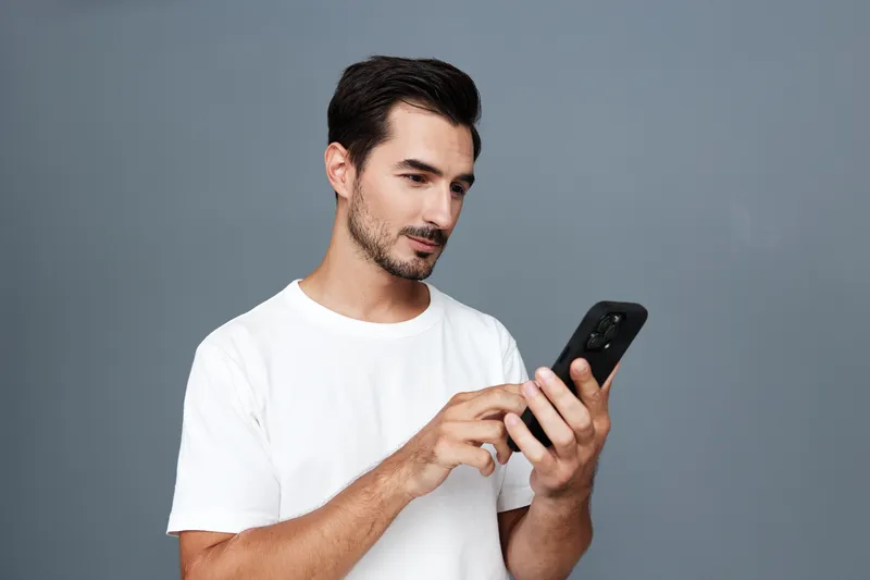Smiling man using mobile cell phone while standing against a plain gray background. Casual style, modern technology, everyday life concept.