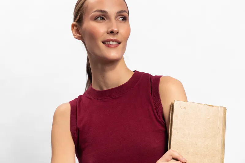 Young woman in sleeveless red top holds a wrapped book, smiling and looking away. Portrait on white background with casual, confident, and positive mood concept.