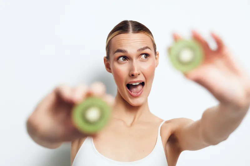 Young woman holds two halves of fresh kiwi fruit close to camera looks excited and happy wearing white tank top isolated on white background with bright natural light