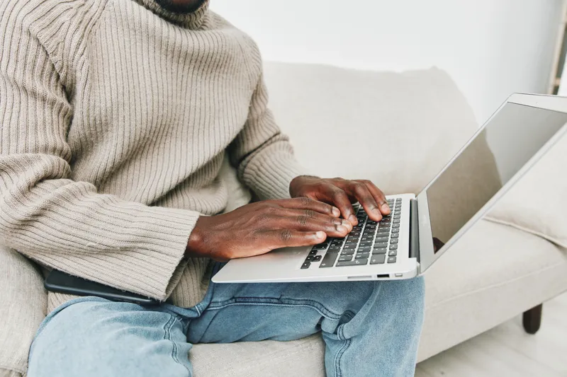 A young Black man working on a laptop while seated on a cozy couch, wearing a beige sweater and casual jeans, depicting a relaxed and productive home environment