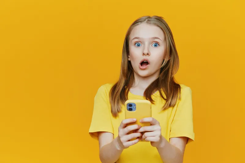 Surprised young girl with smartphone in bright yellow t shirt against a yellow background, expressing shock and excitement, ideal for social media and digital marketing