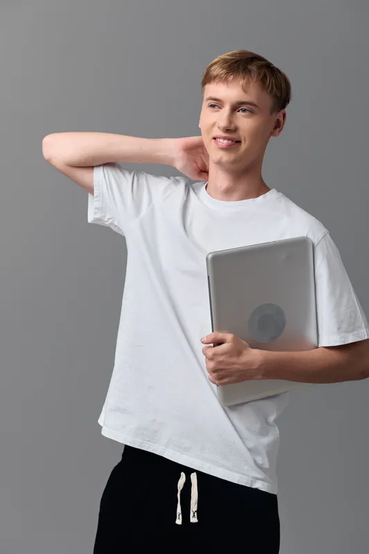 Young man with laptop in casual clothing smiles and stretches in a studio, conveying confidence, modern education and digital technology readiness