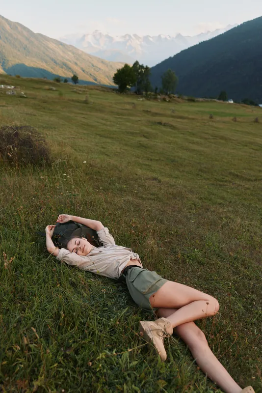 Young woman lies on a grassy hillside in a mountain valley, relaxing and enjoying nature with warm sunlight, casual outfit and a peaceful mood, soft breeze rustling grass as distant peaks glow.