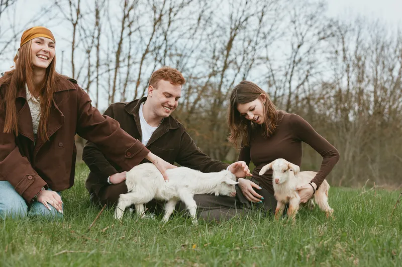 Smiling friends outdoors with two young goats on a grassy field, playful interaction and gentle petting. Natural light, casual clothing, warm mood, rural lifestyle, bonding and kindness.