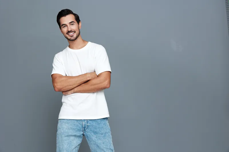 Full body smiling happy cheerful young man dressed in casual white t-shirt and blue jeans, standing confidently with arms crossed against plain gray background.