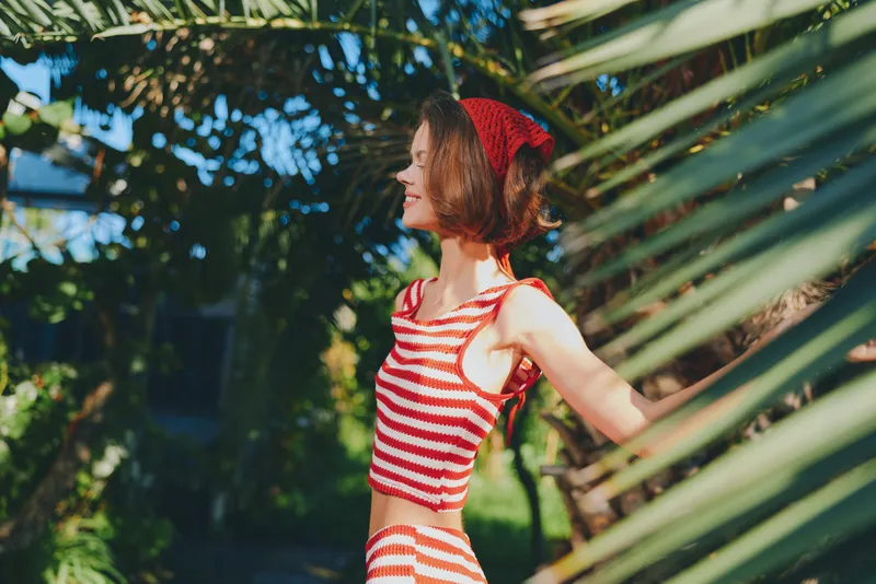 A cheerful young woman in a red striped outfit smiles outdoors among lush greenery, enjoying sunlight and a carefree moment in a vibrant garden setting.