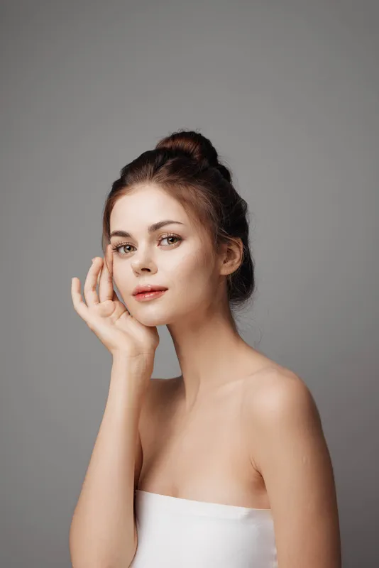 Молодая женщина в белом платье позирует, положив руку на лицо, и смотрит в камеру на сером фоне