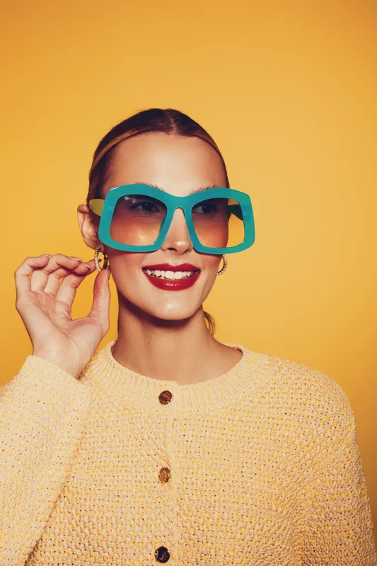 Smiling woman in oversized blue sunglasses and yellow sweater poses against yellow background. Fashion portrait with bright colors and trendy accessories for style concept.