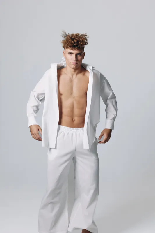 Young man with curly hair wearing white pants and an open white shirt showing toned abs standing against a plain background in a studio setting.