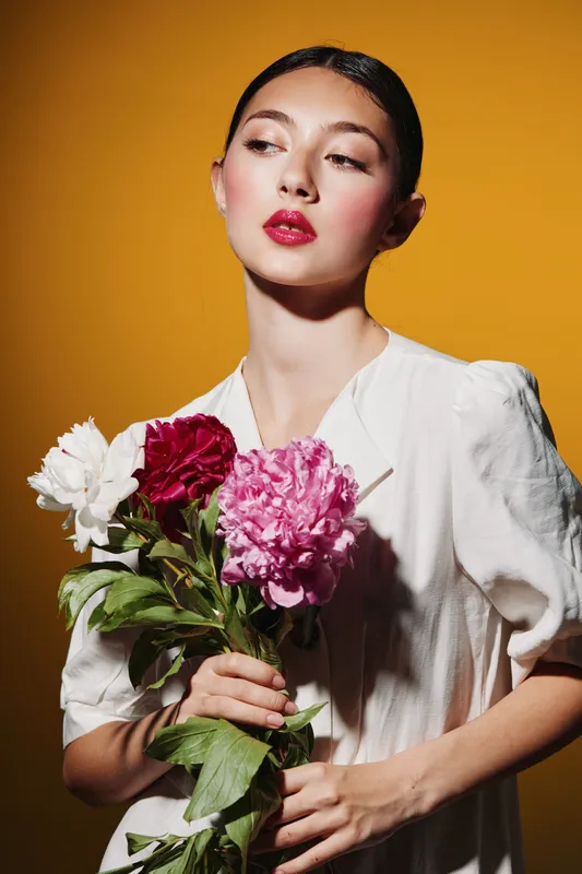 Stylish woman with red lipstick and sleek hair holds bright peony flowers against yellow background. Soft shadows highlight skin texture and colorful beauty fashion concept.