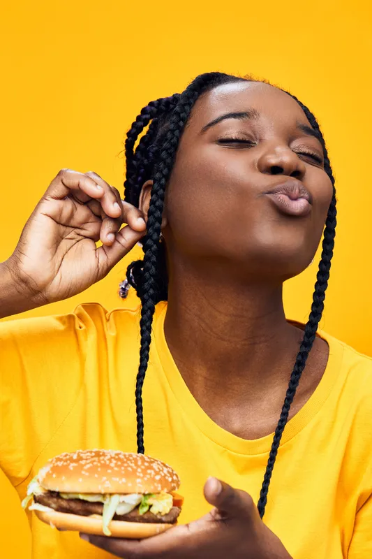Happy woman enjoying a tasty burger against a vibrant yellow background showcasing her love for food and good vibes, ideal for healthy eating themes