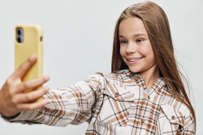 Girl smiling and taking selfie with smartphone, wearing plaid shirt on white background. Portrait of happy teenager with long hair using mobile phone for photo or video call.