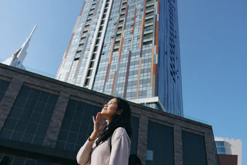 Woman gazes upward beside a towering building, urban exterior architecture under bright daylight, sharing a moment of wonder and awe in a modern cityscape