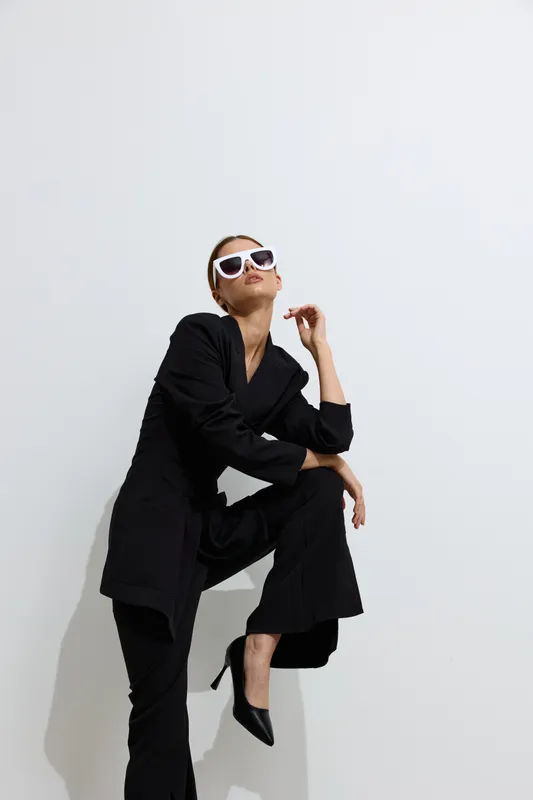 fashion woman wearing black outfit and white sunglasses poses confidently against white background, showcasing modern style and strong attitude in minimalistic setting.