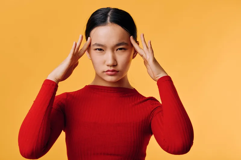 Woman in red sweater with hands on temples, expressing headache or stress with serious face on yellow background. Studio portrait of young Asian female in casual clothing, isolated.