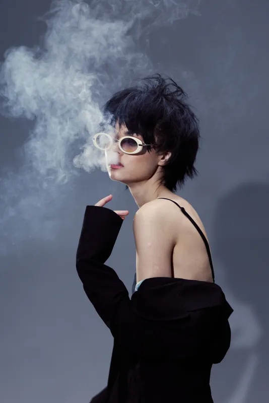 Woman smoking with stylish sunglasses and casual black outfit emitting smoke rings on gray background