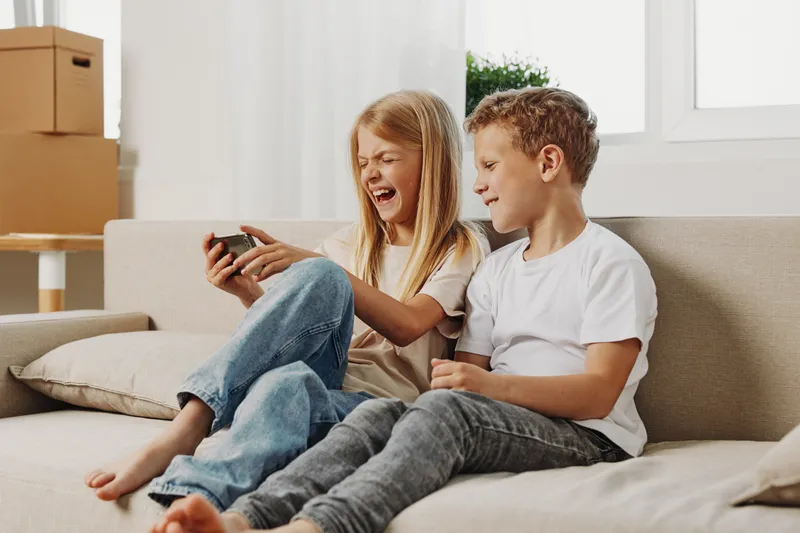 Two children sitting on a couch playing video games together, laughing and enjoying leisure time in a bright living room with natural light. Concept of childhood, fun, and technology.