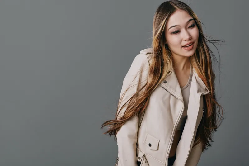 Young woman with long hair wearing beige jacket smiling slightly posing on gray background. Portrait of attractive female model with casual fashionable style and relaxed expression indoors.