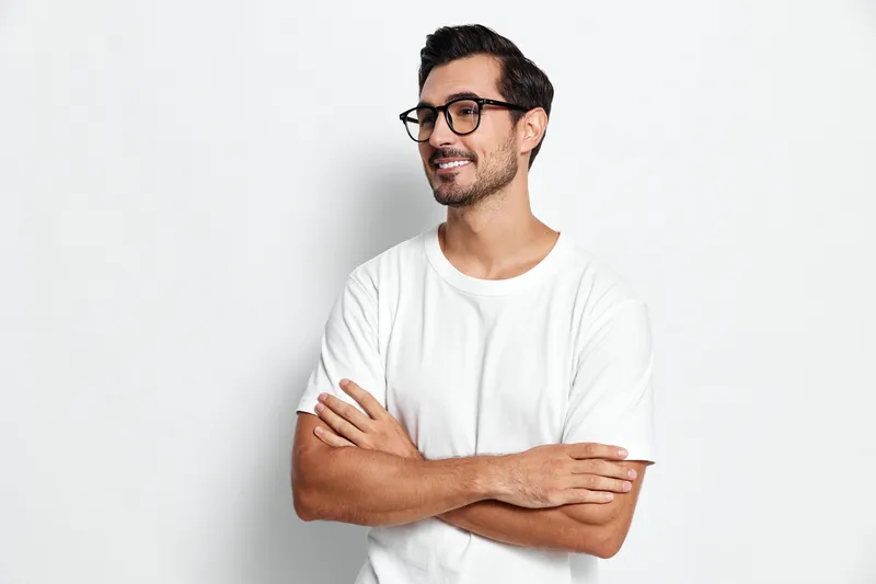 Smiling man wearing glasses standing with arms crossed, looking confidently at the camera, casual style, isolated on a plain light background. Portrait concept.