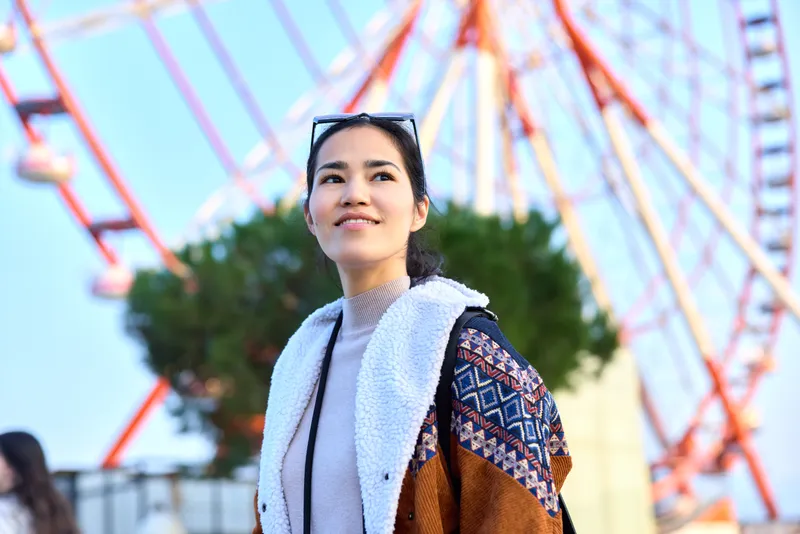 Woman outdoor amusement ferris wheel smiling fashion casual, scarf sunglasses, sunny backdrop, park atmosphere, colorful rides, joyful mood, candid travel vibe.