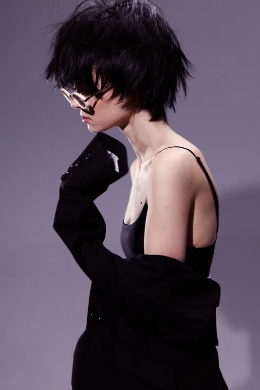 Stylish woman with short black hair wearing sunglasses and dark clothing in a minimalistic studio setting.