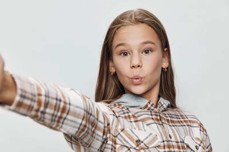 Girl, portrait and playful with plaid shirt making a funny face for selfie. Child with long hair and casual style on plain background for youth and happiness.