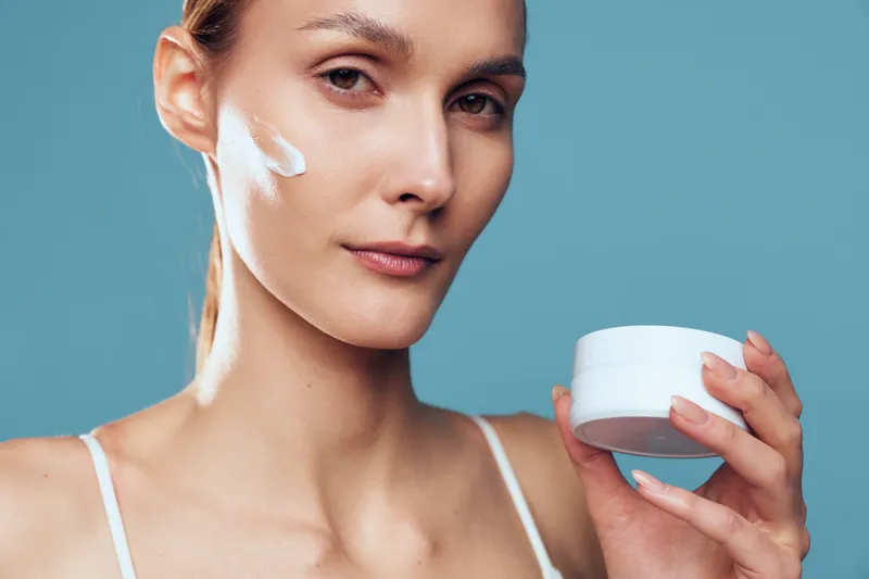 Woman applying white face cream, holding a small jar, close up on blue background, skincare and beauty routine concept, serene expression, soft studio lighting, calm wellness, self care