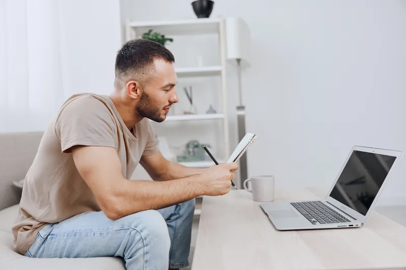Focused man working on a laptop while taking notes, dressed in casual attire. Bright workspace reflecting modern trends and productivity lifestyle.