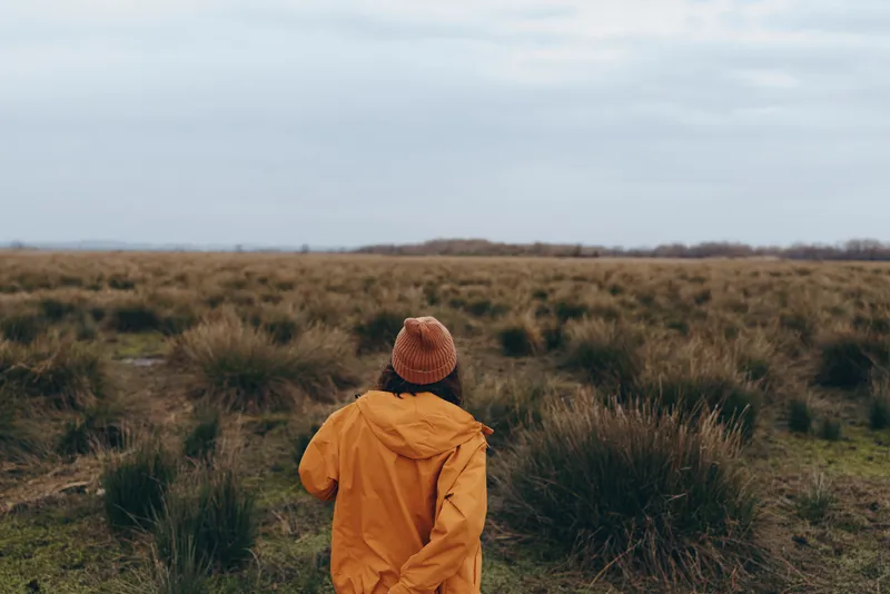 Woman lifestyle, seen from behind, walks outdoors through open grass field, wearing brown jacket and knit hat, cloudy sky above, peaceful nature, solitude, fresh air