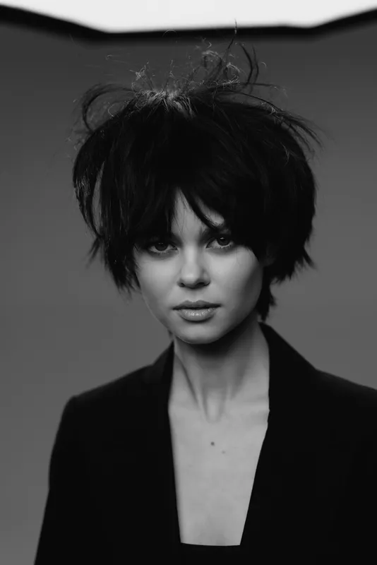 Black and white portrait of a woman with messy hair and intense gaze, dressed in a dark blazer, conveying a moody and artistic style.