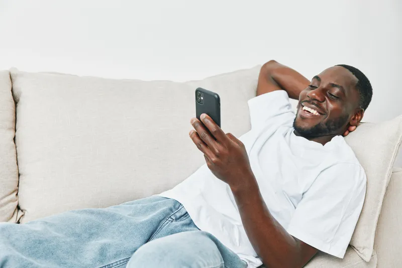 Smiling African man relaxing on a sofa while using a smartphone, wearing casual white t shirt and blue jeans, joyful expression in a bright living room