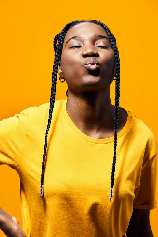 A joyful young Black woman playfully blowing a kiss against a vibrant yellow background, showcasing her stylish braided hair and bright casual outfit