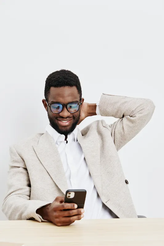 Smiling man using smartphone while sitting at a desk, wearing glasses and a beige blazer, on plain white background. Modern, casual, technology, professional, male, lifestyle.