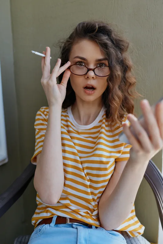 Casual woman with striped shirt sits indoors, wearing glasses, holding a cigarette, surprised expression, cozy home lifestyle moment, intimate daily portrait.