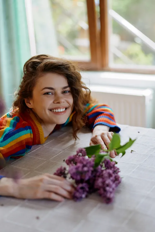 Smiling woman with curly hair sits at a table indoors, holding a bouquet of purple flowers. Natural light from a window creates a warm, cozy home mood, emphasizing happiness and gentle self care.