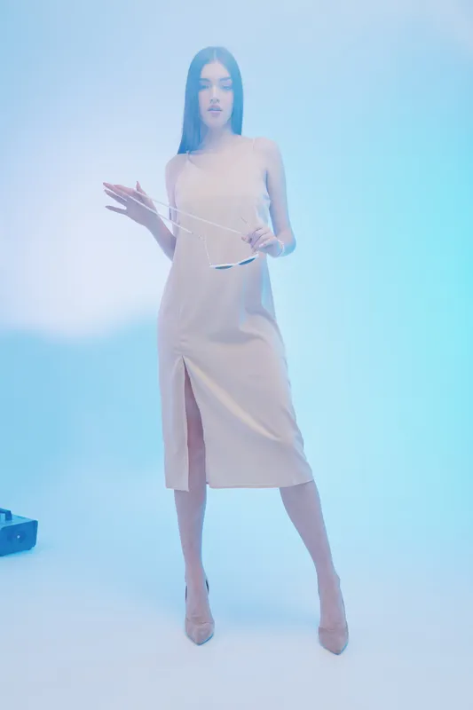 Young woman in a beige sleeveless dress stands confidently holding sunglasses in a soft blue studio light. Concept of beauty, fashion, elegance, and modern style.