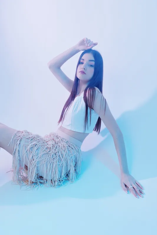 Young woman in stylish fashion outfit poses sitting on the floor in soft blue light. Her long hair and confident expression highlight beauty and modern style concept.