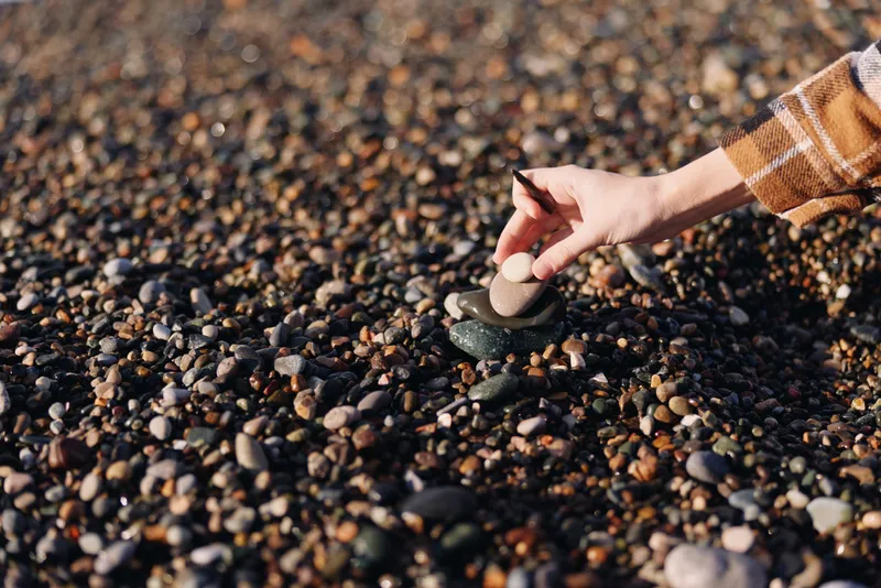 Closeup of a hand picking a smooth stone among pebbles on a sunlit beach, showcasing natural texture and detail, outdoor scene with calm coastal atmosphere and tactile surface