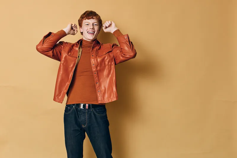 Young man with red hair smiling and raising hands excitedly in brown jacket and orange sweater standing against beige background in casual style with jeans and belt.